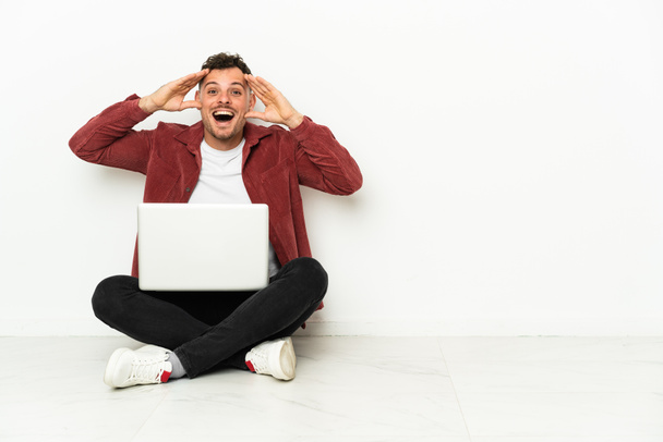 Young handsome caucasian man sit-in on the floor with laptop with surprise expression - Photo, Image