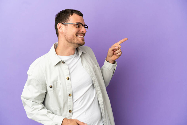 Brazilian man over isolated purple background pointing finger to the side and presenting a product - Photo, Image