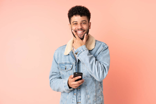 Young Moroccan man using mobile phone isolated on pink background happy and smiling - 写真・画像