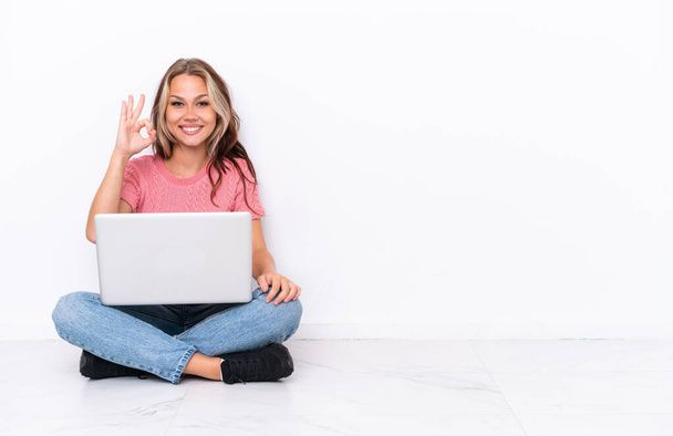 Joven chica rusa con un ordenador portátil sentado en el suelo aislado sobre fondo blanco que muestra signo de ok con los dedos - Foto, Imagen