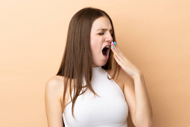 Молода біла жінка ізольована на бежевому фоні позіхання і покриває широкий відкритий рот рукою
 - Фото, зображення