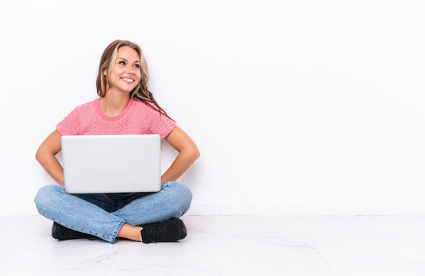 Joven chica rusa con un portátil sentado en el suelo aislado sobre fondo blanco posando con los brazos en la cadera y sonriendo - Foto, imagen