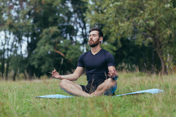 A young man with a beard sitting in the park on a mat meditates, performs exercises to improve breathing, active lifestyle - Фото, изображение