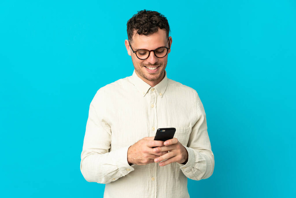 Young caucasian handsome man isolated on blue background sending a message with the mobile - Фото, зображення