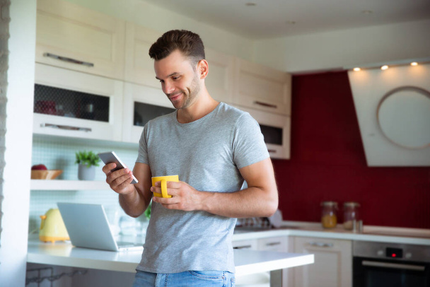 Handsome confident modern man in gray t-shirt stands with phone in hands and drinks coffee from the yellow cup on the kitchen at home - Photo, image
