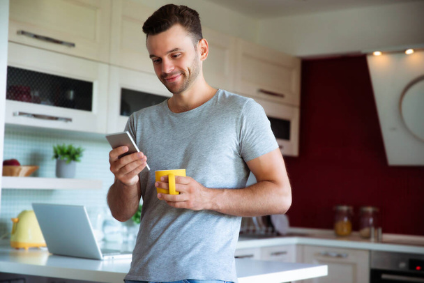 Handsome confident modern man in gray t-shirt stands with phone in hands and drinks coffee from the yellow cup on the kitchen at home - Фото, зображення