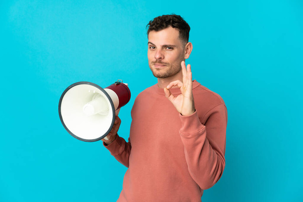 Young caucasian handsome man isolated on blue background holding a megaphone and showing ok sign with fingers - Photo, Image