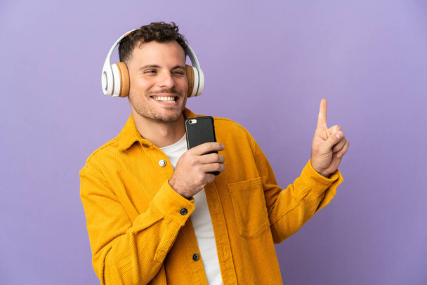 Mladý kavkazský pohledný muž izolovaný na purpurovém pozadí poslech hudby s mobilní a zpěv - Fotografie, Obrázek