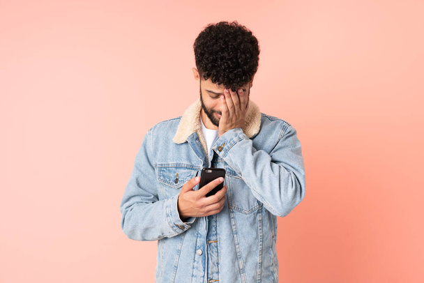 Hombre joven marroquí usando teléfono móvil aislado sobre fondo rosa con expresión cansada y enferma - Foto, Imagen
