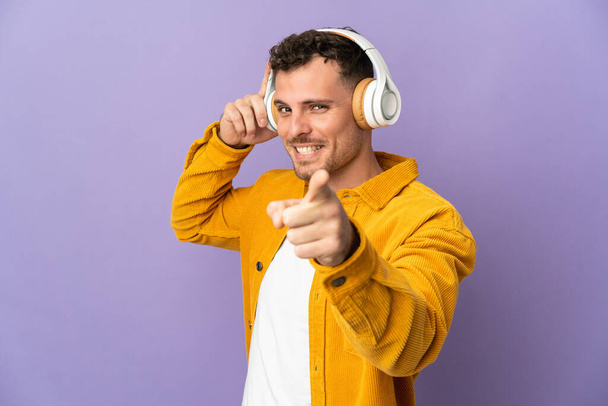 Jonge blanke knappe man geïsoleerd op paarse achtergrond luisteren muziek en wijzen naar het front - Foto, afbeelding
