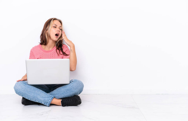 Junge russische Mädchen mit einem Laptop auf dem Boden sitzend isoliert auf weißem Hintergrund gähnen und weit geöffneten Mund mit der Hand bedecken - Foto, Bild