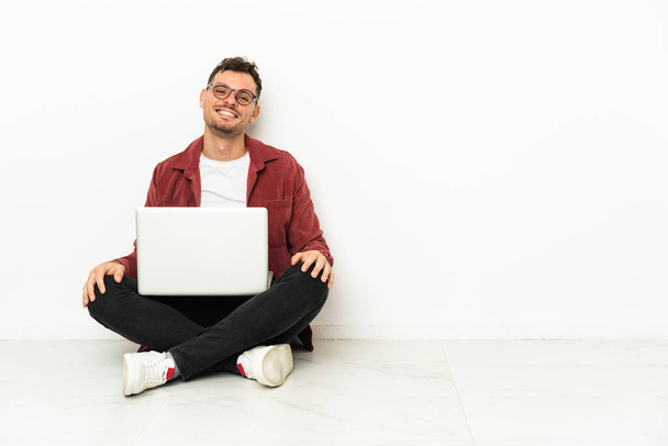 Νεαρός όμορφος καυκάσιος άνδρας κάθεται στο πάτωμα με laptop με γυαλιά και χαρούμενος - Φωτογραφία, εικόνα