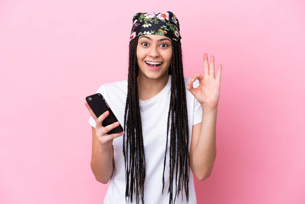 Teini tyttö punokset yli eristetty vaaleanpunainen tausta matkapuhelimella ja tekee OK merkki - Valokuva, kuva