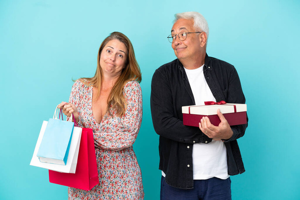 Střední věk pár s nákupní taškou a dárek izolované na modrém pozadí dělat pochybnosti gesto při zvedání ramen - Fotografie, Obrázek