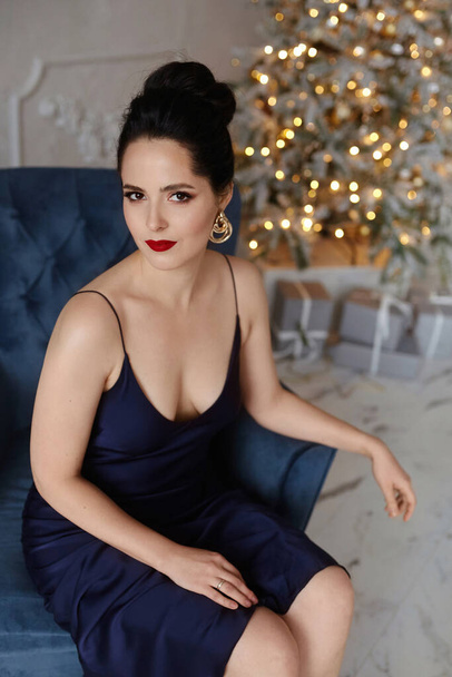 Hermosa chica modelo con maquillaje brillante y peinado modish usando vestido de noche azul oscuro se sienta en un sillón vintage con luces navideñas festivas en el fondo - Foto, Imagen