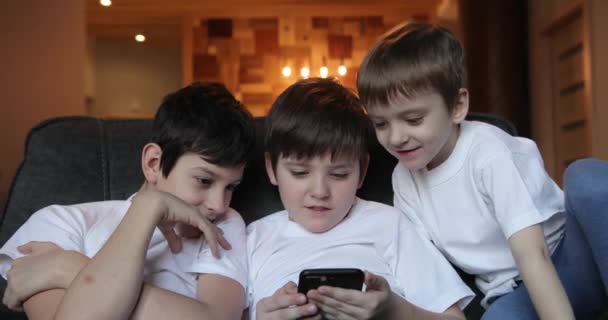 lachende kinderen die grappige video keken op een smartphone die samen op de bank zat. kinderen die graag spelletjes spelen of zich vermaken met mobiele apps op de telefoon thuis. - Video