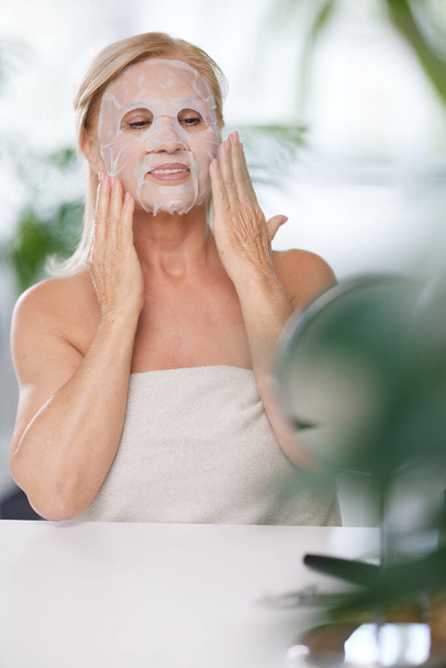 Старшая женщина надевает маску красавицы на лицо, сидя дома, завернутая в полотенце. Старшая женщина с помощью косметики - Фото, изображение