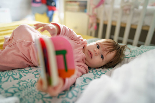 1つの小さな白人の赤ん坊は、明るい日の子供時代に遊んで本のおもちゃを保持し、コンセプトレジャーを成長させるカメラを探して家でベッドの上に一人で横たわっています - 写真・画像