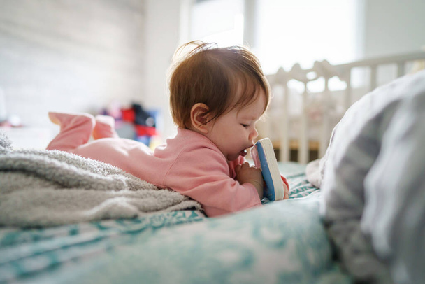 Ein kleines kaukasisches Baby, das zu Hause allein auf dem Bett liegt und in die Kamera blickt, mit Bücherspielzeug in der Hand, das in der Kindheit spielt und in der Freizeit aufwächst - Foto, Bild