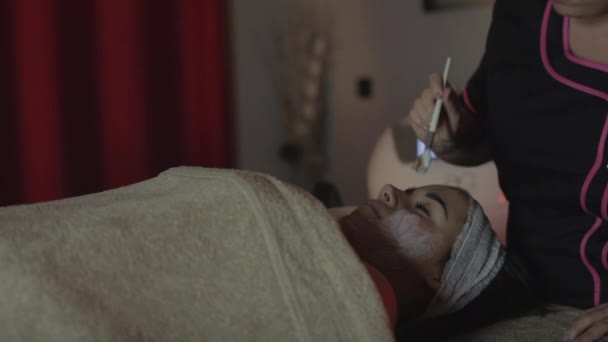 Mujer joven relajándose bajo el toque suave del especialista que aplica en sus mejillas mascarilla facial blanca con efectos rejuvenecedores - Metraje, vídeo