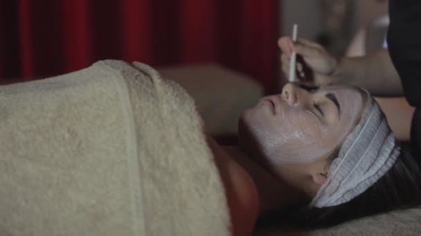 Young woman relaxing under the gentle touch of the specialist applying on her cheeks white facial mask with rejuvenating effects - Footage, Video