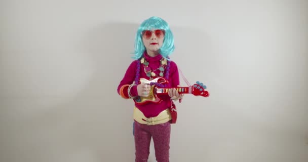 Guitarra adorable divertida del jugador de la muchacha del cabrito feliz y canta en el micrófono que usa el equipo colorido del estallido como gafas de sol y peluca colorida - Imágenes, Vídeo