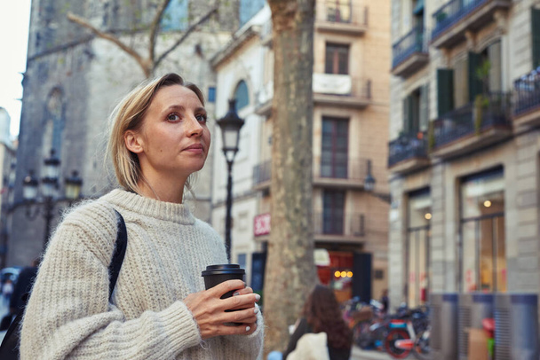 Attractive young caucasian woman looking thoughtful while holding a cup of coffee in her hand in the city during the day - Foto, imagen