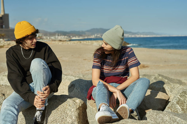 Two riveted young people sitting on rocks beside a beach and having a conversation - Фото, изображение