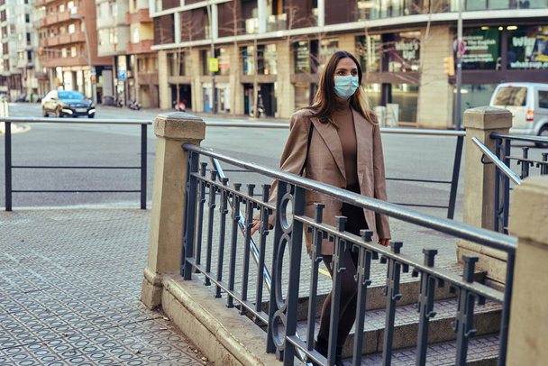 Unsmiling lady gradually walking down stairs while balancing on the railing and wearing a medical mask - Fotoğraf, Görsel