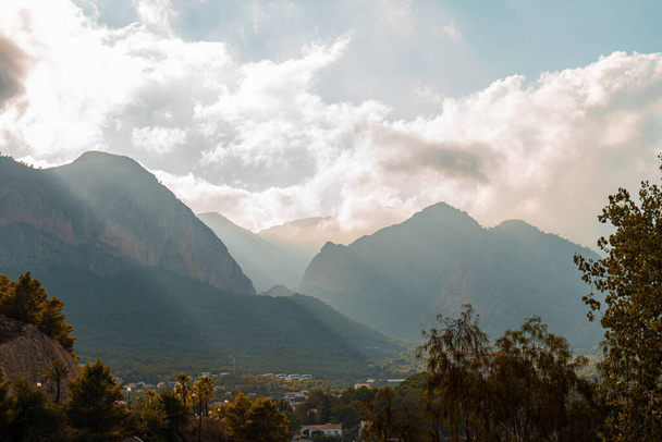 A town surrounded by a mountain range and trees on a sunny, cloudy day - Fotó, kép