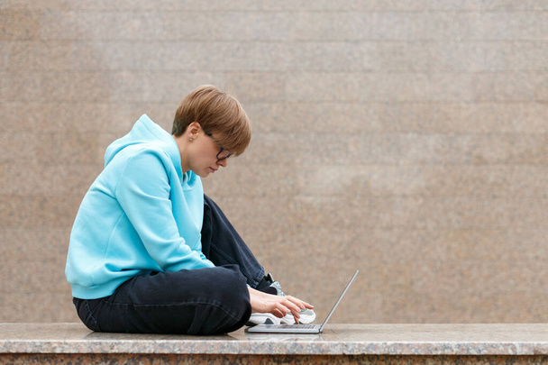 Photo of a young hipster woman with a laptop in an urban environment. She works intently, sitting on the parapet against the granite wall. - Photo, Image
