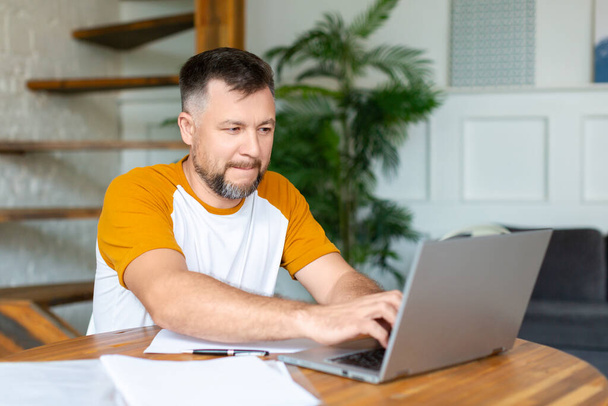 Ein Mann mittleren Alters arbeitet zu Hause vor einem Laptop-Monitor. Er tippt Text. - Foto, Bild