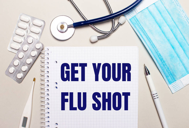 On a light gray background, a light blue disposable face mask, a stethoscope, an electronic thermometer, pills, a pen and a notebook with the inscription GET YOUR FLU SHOT. Medical concept - Fotó, kép