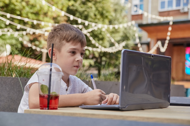 kleine schooljongen doet huiswerk buiten met laptop, het schrijven van notities in copybook - Foto, afbeelding