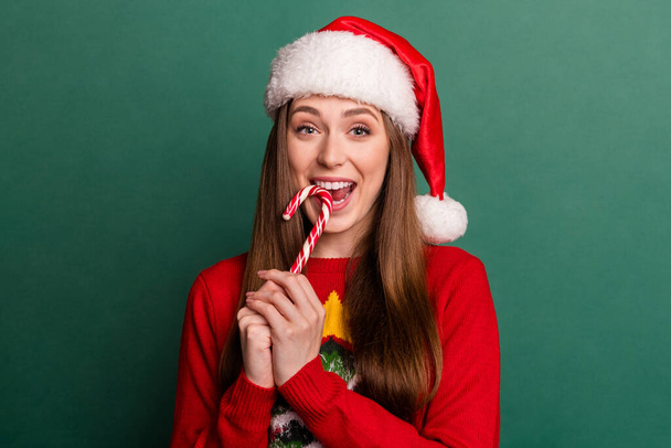 Photo of young woman happy positive smile eat candy yummy wear santa hat isolated over green color background - Valokuva, kuva