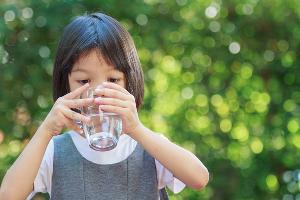 Portrait of a 4 to 6 year old Thai kid Asian girl with cute face holding a glass of water. She is drinking water to refresh her body. The background is green. Concept of girl drinking water - Photo, image