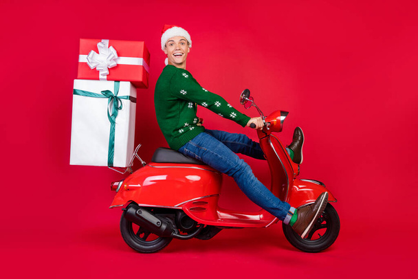 Full length body size photo man smiling guy driving bike with gifts on wearing festive cap isolated vivid red color background - Fotó, kép