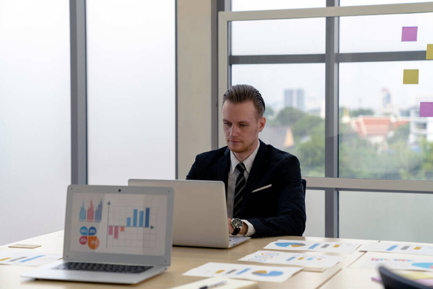 Zeki, yakışıklı, beyaz iş adamı laptopla modern ofiste çalışma masasına güvenerek odaklanıyor. Ekranda grafikler, diyagramlar ve çizelgeler olan tablet bilgisayar. finansal analiz. - Fotoğraf, Görsel