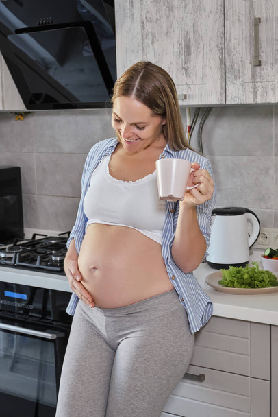 Выстрел симпатичной женщины держит руку на животе, пьет горячий чай, держит кружку с напитком, позирует против кухни интерьера в одиночку, наслаждается спокойной атмосферой, будучи беременной. Исключая мать дома
 - Фото, изображение