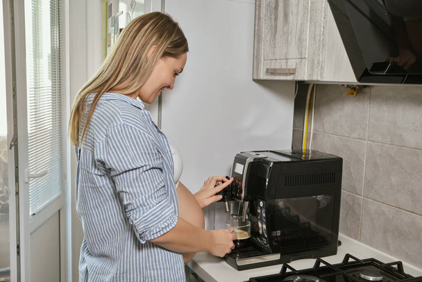 Genç hamile kadın mutfakta kahve içiyor, kahve makinesi kullanıyor. - Fotoğraf, Görsel