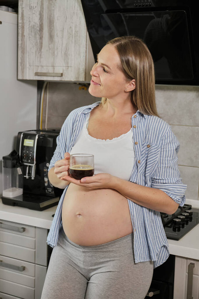 Знімок приємного вигляду жінки тримає руку на животику, п'є гарячий чай, тримає кухоль з напоями, позує проти кухонного інтер'єру, насолоджується спокійною атмосферою, вагітніє. Розшукуючи маму вдома
 - Фото, зображення