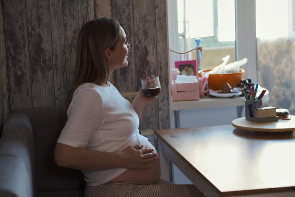 Der neue Tag beginnt mit frischem Kaffee. Schöne fröhliche schwangere Frau hält Kaffeetasse und schaut lächelnd weg, während sie zu Hause in der Küche sitzt - Foto, Bild