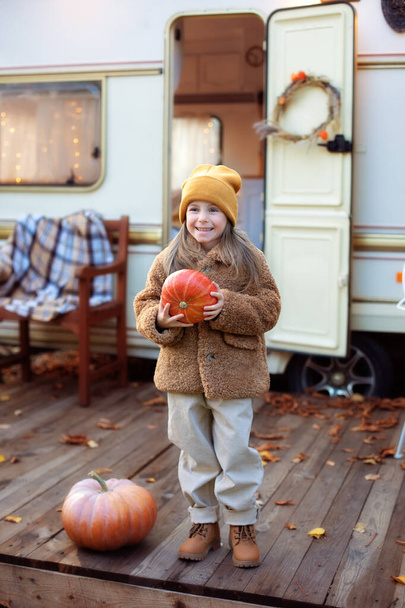 Happy Halloween. Smiling girl standing on the porch with a pumpkin in her hands. Trick-or-treat. Happy little girl playing near camping at RV home in the garden. Child in cozy campsite fall backyard.  - Valokuva, kuva