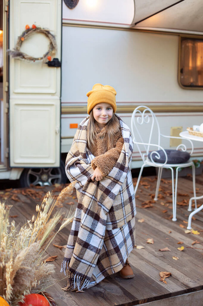 Smiling young girl with warm blanket standing on porch RV house in fall garden. Child covered with blanket on terrace house to warm up. Child girl in campsite backyard. Concept camping, adventure  - Foto, Bild