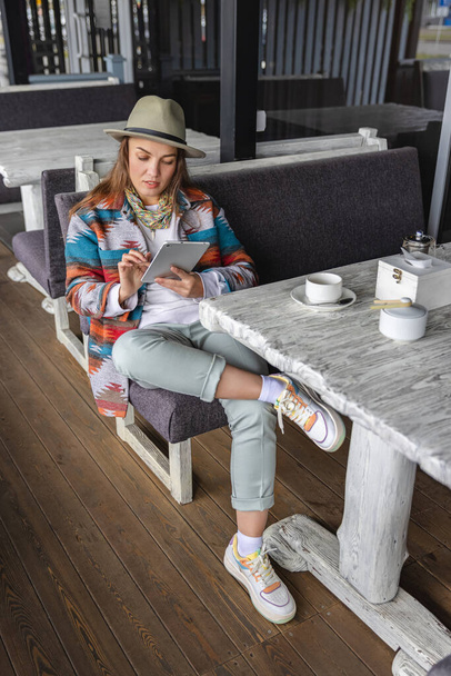 An attractive woman in a hat and a bright shirt, sits in a cafe, chatting in a tablet. The brown-haired woman spends time in autumn, spring, cold weather, fresh air. - Photo, image