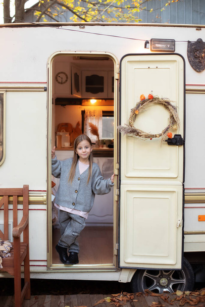 Smiling little girl in casual clothes standing on porch RV house in garden. Cute young girl stand near trailer door. Child in cozy campsite fall backyard. Concept camping, outdoor, nature, adventure  - 写真・画像