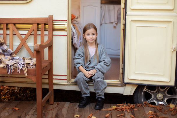 Smiling little girl in casual clothes sitting on porch RV house in garden. Cute young girl sitting near trailer door. Child in cozy campsite fall backyard. Concept camping, outdoor, nature, adventure - Valokuva, kuva