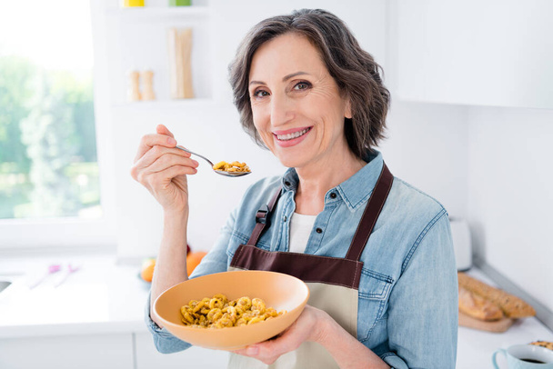 Photo of cheerful positive joyful old woman hold hand plate spoon cereal indoors inside house home kitchen - Foto, Imagem