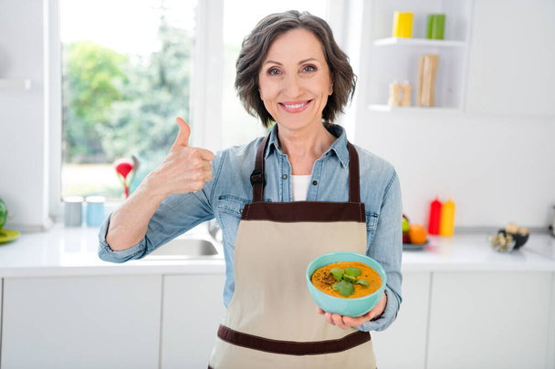 Photo of age funny lady hold soup show thumb up wear apron jeans shirt at kitchen alone - Fotografie, Obrázek
