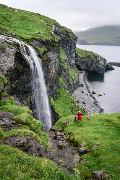 赤いジャケットの旅行者は、ヴァガール島、フェロー諸島のSkardsafossur滝を写真に収めています - 写真・画像